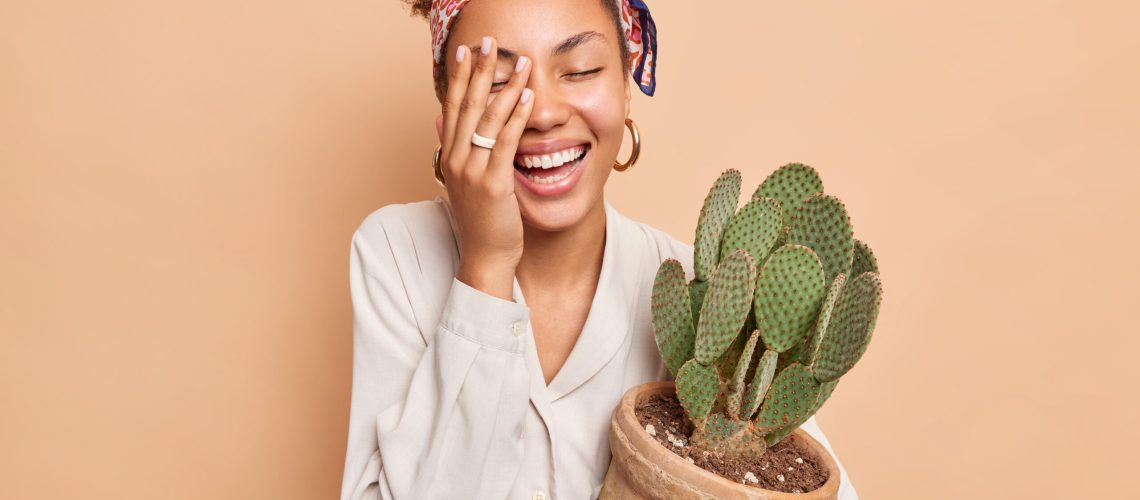 Cheerful dark skinned female model makes face palm smiles broadly has happy mood embraces pot with cactus wears white shirt and headkerchief isolated over beige background takes care of houseplants